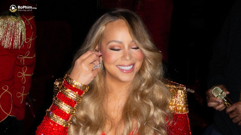 Mariah Carey mặc trang phục đỏ lấp lánh, cười rạng rỡ trong hậu trường với ánh đèn sân khấu phía sau.