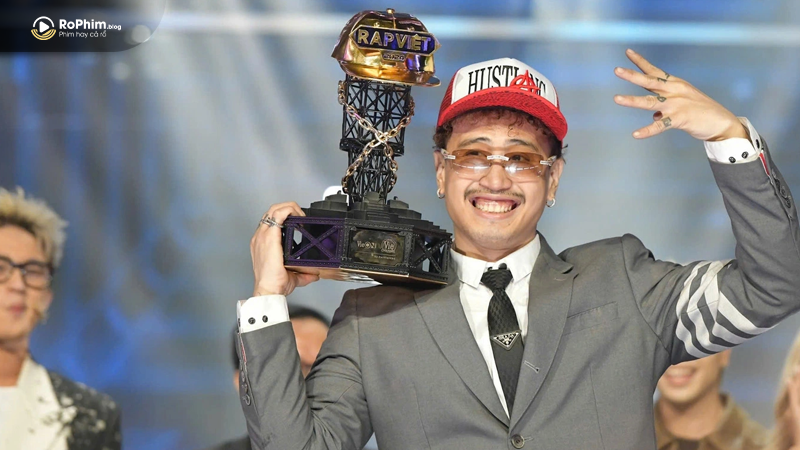 Robber giơ cao chiếc cúp vô địch Rap Việt 2024, nở nụ cười rạng rỡ trong bộ vest xám và mũ đỏ nổi bật.