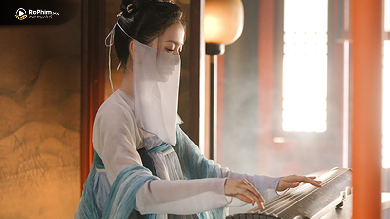 Người phụ nữ đeo mạng che mặt, mặc y phục xanh, đang chơi đàn trong ánh sáng dịu